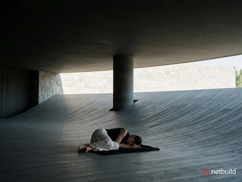 Cinematic wide shot of a solitary barefoot figure curled in a tight fetal position on a sweeping, concave floor of grey wooden planks. The person wear...